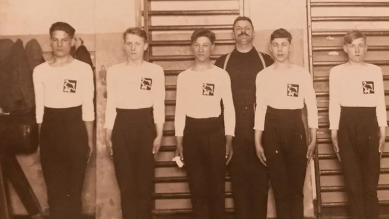 Groep in sportkleding met arend-emblemen, voor wandrekken in een gymnastiekzaal.